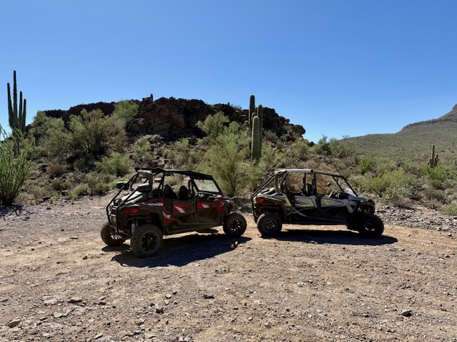 Two side-by-side UTVs parked in a desert landscape surrounded by dirt and cacti